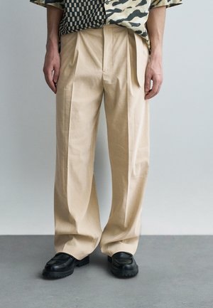 Pantaloni beige a gamba larga plissettati abbinati a mocassini neri con suola spessa e camicia fantasia.