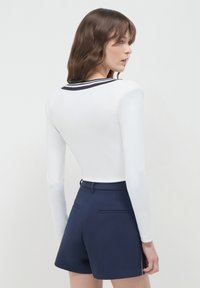 Frau, die sich seitlich dreht und ein weißes Langarmoberteil mit schwarzem gestreiftem Kragen und navyblauen, hoch geschnittenen Shorts trägt, vor einem schlichten Hintergrund.