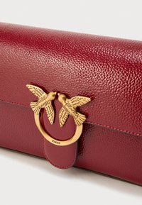 Τσάντα χειρός από δέρμα μπορντό με υφή, με κούμπωμα σε χρυσό τόνο που έχει σχήμα δύο πουλιών. Το όνομα της μάρκας "PINKO" εμφανίζεται στα μεταλλικά μέρη.