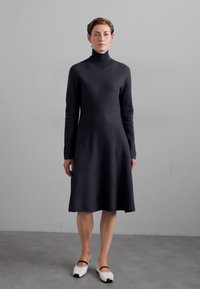 Schwarzes Rollkragenkleid mit langen Ärmeln, tailliertem Oberteil und ausgestelltem Rock. Kombiniert mit weißen flachen Schuhen mit schwarzen Zehenakzenten.