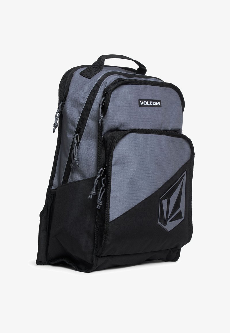 Sac à dos Volcom gris et noir avec plusieurs compartiments zippés, une poignée supérieure et un écusson logo à l'avant.