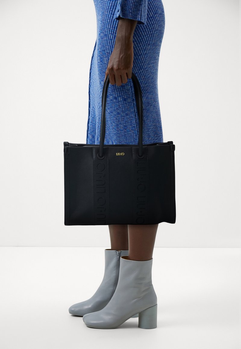 Personne portant une robe côtelée bleue et des bottines gris clair, tenant un sac cabas noir avec doubles anses et logo doré.