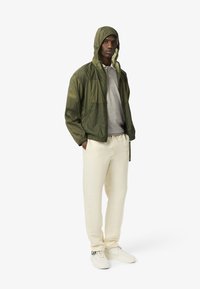 Grön lätt vikbar jacka med huva, randig tröja, krämfärgade sweatpants och vita sneakers. Visad på en stående manlig figur.