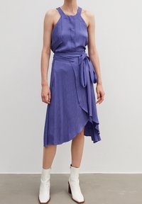 Lila klänning med ett texturerat vågmönster, halterneck och knytmidja, med en asymmetrisk fåll. Bärs med vita anklar boots.