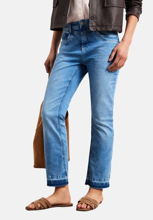 Femme portant un jean bleu clair avec des ourlets effilochés, une veste en cuir marron, des sandales beiges et tenant un sac en daim marron à son côté.