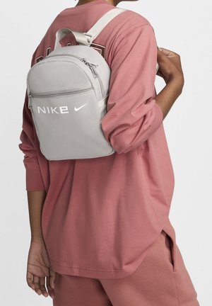 Personne portant un sac à dos Nike gris clair sur une épaule, vêtue d'un haut ample à manches longues rose poussiéreux et d'un pantalon assorti.