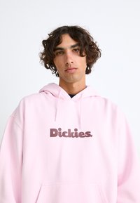 Felpa rosa realizzata in tessuto morbido, con tasca a canguro e logo "Dickies" marrone scuro centrato sul petto.