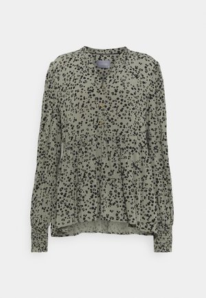 Blouse verte avec un motif à pois noirs, devant boutonné, manches longues avec des poignets froncés, et un design en étages évasé. Texture de tissu douce.