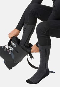 Schwarzer Skistiefel mit metallischem Beschlag, kombiniert mit gerippten dunkelgrauen Socken, die entlang der Seiten ein Logo tragen. Fokus auf Passform und Komfort.