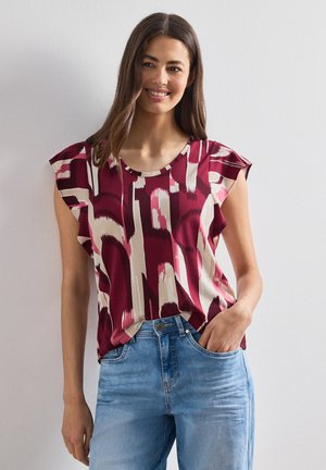Lächelnde Frau, die ein ärmelloses burgunderfarbenes und beiges gemustertes Top mit Rüschenärmeln und hellblaue Jeans trägt, mit der Hand in der Tasche vor einer hellen Wand.