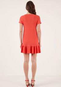 Short-sleeve, coral dress with a ruffled hem, textured material, and a scooped neckline. Model is wearing matching red sandals.