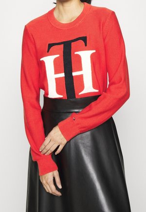 Frau trägt einen roten Pullover mit großen schwarzen und weißen "TH"-Buchstaben und einen schwarzen Lederrock, die Hände sanft verschränkt auf der Taille.