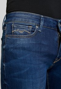 Vaqueros de mezclilla azul oscuro con forma ajustada. Incluye cremallera, cinco bolsillos y costuras en contraste. La tela tiene una textura suave.