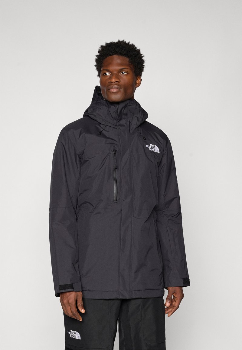 Jachetă impermeabilă neagră cu glugă, fermoar frontal, două buzunare pe piept și manșete ajustabile. Prezintă logo-ul The North Face pe piept.