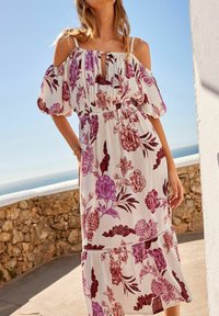 Donna che indossa un vestito bianco a spalle scoperte con stampa floreale in viola e rosso, in piedi all'aperto vicino a un muro di pietra sotto un cielo azzurro e sereno.