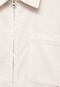 Tissu écru avec tissage diagonal visible, fermeture éclair en haut au centre et une poche rectangulaire cousue sur le côté droit.