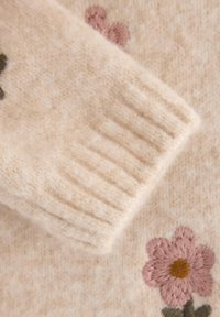 Beige strikket stoff med teksturert ribbestrikket mansjett. Har broderte rosa blomster og grønne blader, som gir detalj til den myke overflaten.