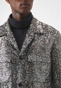 Giacca nera e argento decorata con paillettes, con colletto, chiusura a bottone e taschino sul petto; superficie strutturata che riflette la luce.