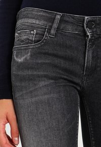 Vaqueros de mezclilla negra con un acabado desgastado, diseño de cinco bolsillos, cierre de botón y una superficie texturizada sutil, con señales de uso cerca de los bolsillos.