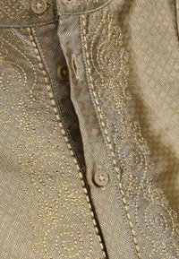 Beige blouse met gestructureerde stof met een geometrisch patroon, geborduurde bloemenaccenten en knoopsluitingen aan de voorkant.