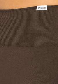 Bruine ribstof met een gladde textuur, voorzien van een opvallend taillebandlabel met de tekst "SMILODOX." Het algehele ontwerp is eenvoudig en aansluitend.