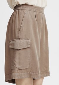 Shorts beige, arrivant aux genoux, fabriqués en tissu léger. Dotés d'une taille élastique et d'une poche latérale avec rabat. Texture lisse et coupe décontractée.