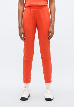 Pantaloni sportivi arancione brillante con cintura elastica, taglio slim e dettagli riflettenti, abbinati a sneakers bianche e calzini corte.