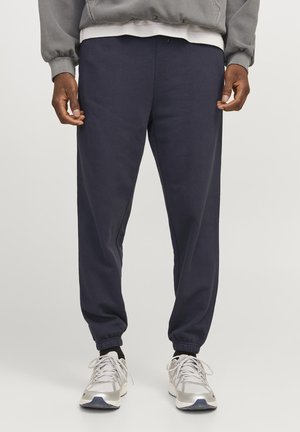Homme portant un pantalon de jogging bleu marine foncé avec des poignets élastiques, un sweat-shirt gris, des chaussettes noires et des baskets blanches et argentées, debout sur un fond uni.