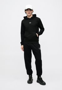 Joven de pie con sudadera con capucha negra EA7, pantalones deportivos negros a juego, zapatillas negras y una gorra de béisbol blanca, sobre un fondo claro liso.