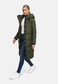 Cappotto puffer verde oliva con zip e cappuccio, caratterizzato da un design a quadri e grandi segmenti. Abbinato a jeans blu scuro e sneakers bianche.