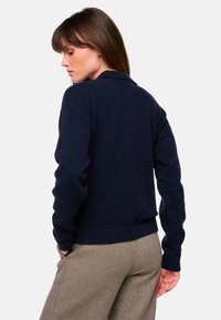 Marineblauer Pullover mit langen Ärmeln und gerippten Bündchen. Das Design verfügt über einen Kragen und einen taillierten Bund, kombiniert mit beigen strukturierten Hosen.