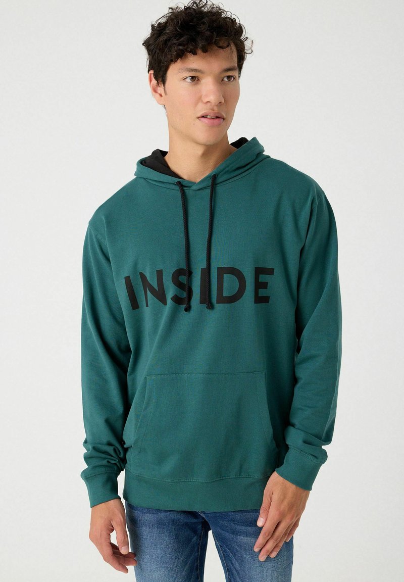 Sudadera verde con gran texto negro "INSIDE", bolsillo canguro, capucha con cordón y puños y dobladillo acanalados. Combinada con jeans azules.