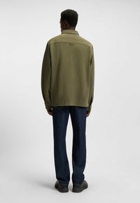 Olive green, ribbed long-sleeve shirt with a collar and buttoned cuffs, paired with dark blue straight-leg jeans and black shoes.