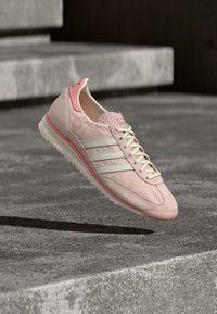 Zapatilla deportiva de color rosa claro con materiales de gamuza y malla, tres rayas blancas y un diseño texturizado en el lateral. Suela delgada y curva.