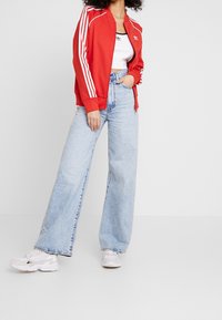 Červená zipová bunda s bílými pruhy, spárovaná s černým crop topem a světle modrými širokými džíny. Koutfitu dominují bílé tenisky.