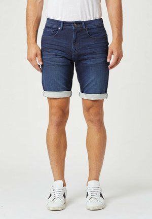 Shorts en denim bleu foncé, avec un design à revers, poches avant et fermeture éclair. Portés avec des baskets blanches.