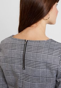 Mustavalkoinen ruudullinen mekko, jossa on näkyvä vetoketju selässä, rakenteellinen kaula-aukko ja teksturoitu kangas.