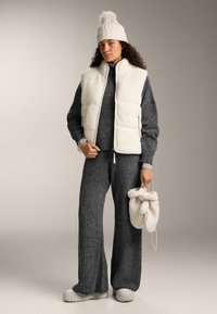 Frau trägt eine cremefarbene Strickmütze mit Bommel, eine cremefarbene Steppweste, einen anthrazitfarbenen Strickpullover und Hose und hält cremefarbene flauschige Fäustlinge mit Handgelenkriemen.