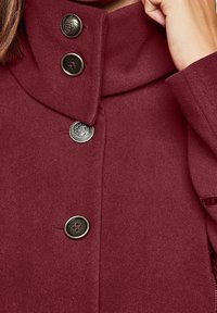 Abrigo de lana burdeos con un cuello alto, que presenta dos botones redondos en tono plata y un acabado texturizado. Tejido suave con detalles en hardware.