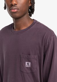 Long-sleeve, crew-neck, purple t-shirt made of soft cotton with a small chest pocket and logo patch. Features stitched hems and relaxed fit.