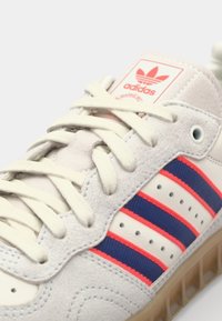 Biele tenisky z semišových a kožených materiálov, s červenými a modrými prúžkami, svetlými šnúrkami a textúrovanou gumovou podrážkou.