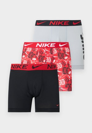 Atletický muž stojaci naboso, oblečený v čiernych boxerkách Nike s červeným pásom, pozerá dolu a mierne do strany na jednofarebnom pozadí.