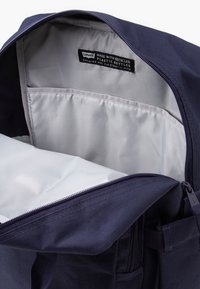 Jūrinės mėlynos spalvos kuprinė su lygia išore, pilka vidine apdaila ir etikete, nurodančia perdirbtas medžiagas. Uždarymas su užtrauktuku ir šoninės kišenės detalė.