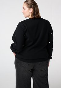 Zwarte sweater met hartvormige versieringen op de mouwen, met een relaxte pasvorm en zachte stof. Gecombineerd met donkergrijze jeans met wijde pijpen.