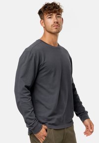Graues Langarmshirt mit strukturiertem Muster, Rundhalsausschnitt und gerippten Bündchen, kombiniert mit olivgrünen Hosen, die Hände in den Taschen.