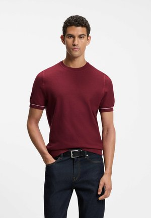 Hombre con cabello rizado, vestido con un suéter ajustado de manga corta color granate y jeans azul oscuro, de pie con una mano en el bolsillo.