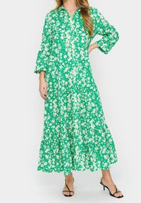 Grön blommig klänning med långa ärmar, figursydd överdel, lagd kjol och en krage. Materialet verkar lätt och har en mjuk textur.