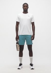 Άνδρας που στέκεται φορώντας λευκό T-shirt Puma, τυρκουάζ σορτς Puma, λευκές κάλτσες, αθλητικά παπούτσια, κρατώντας μπεζ παγούρι Puma στο δεξί χέρι.