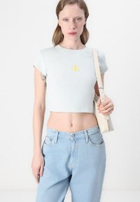 Světle modré zkrácené tričko vyrobené z ribstop látky s žlutým logem, spárované s džínami s vysokým pasem v světle modré barvě a krémovou kabelkou přes rameno.