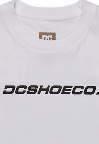 Bílý bavlněný tričko s výrazným černým potiskem "DCSHOECO." na hrudi. Jednoduchý kulatý výstřih. Hladká textura, standardní střih.